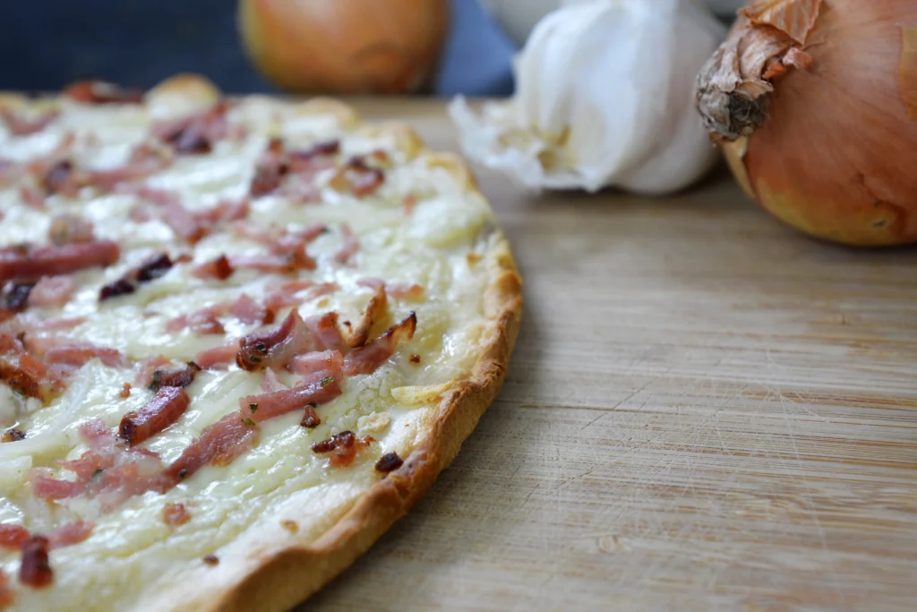 Photo d’une Flammekueche maison dorée et croustillante, garnie d’oignons, de crème fraiche et de lardons