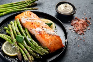 Pavé de saumon grillé avec des asperges vertes et une crème à l’ail, servi avec des tranches de citron sur une assiette noire – Recette gourmande et saine