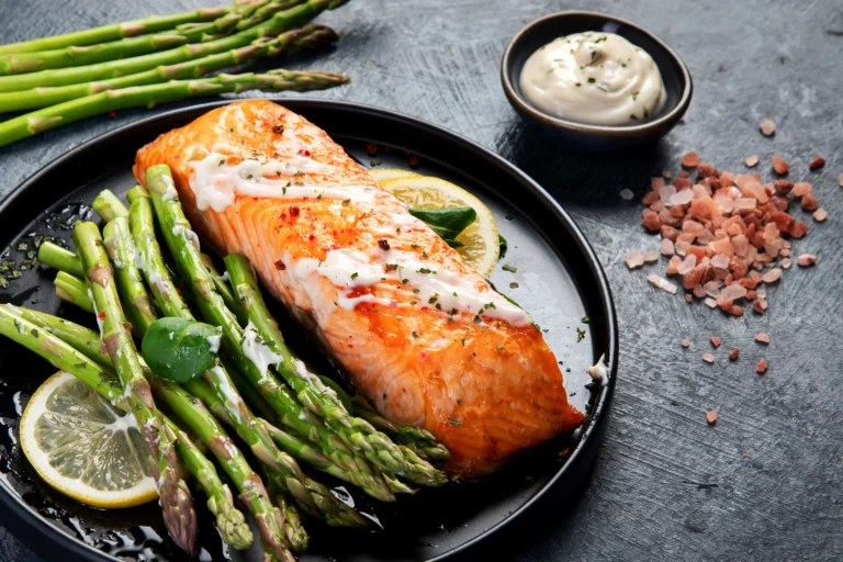 Pavé de saumon grillé avec des asperges vertes et une crème à l’ail, servi avec des tranches de citron sur une assiette noire – Recette gourmande et saine