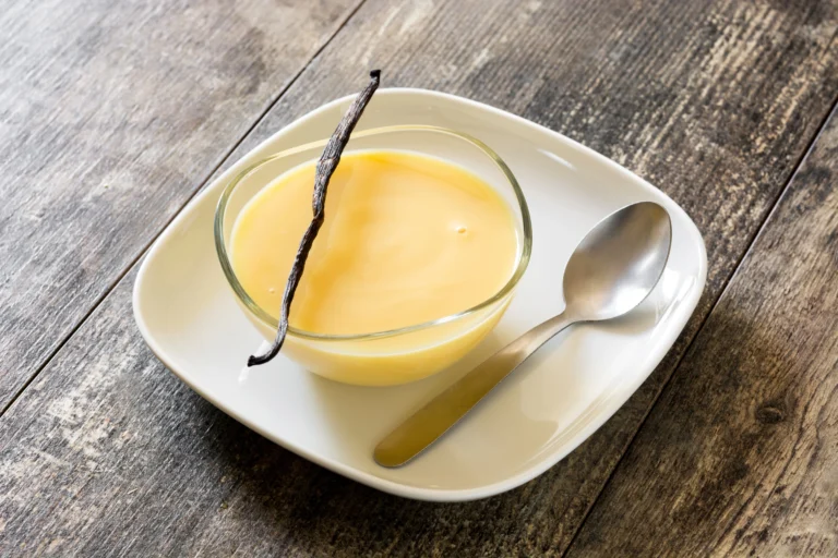 Crème anglaise maison servie dans un petit bol blanc, nappée légèrement sur une cuillère, avec une gousse de vanille en décor