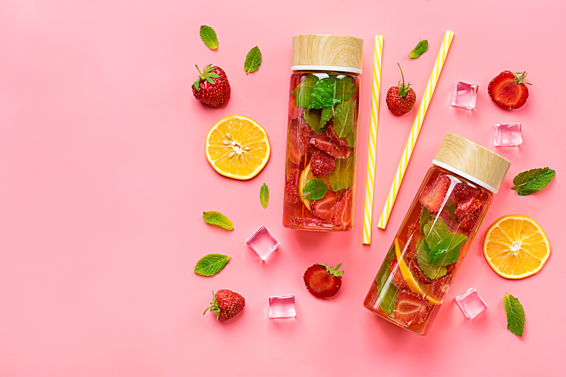 Deux bouteilles d’eau aromatisée avec des tranches de fraise, d’orange et des feuilles de menthe sur fond rose, accompagnées de glaçons et pailles colorées