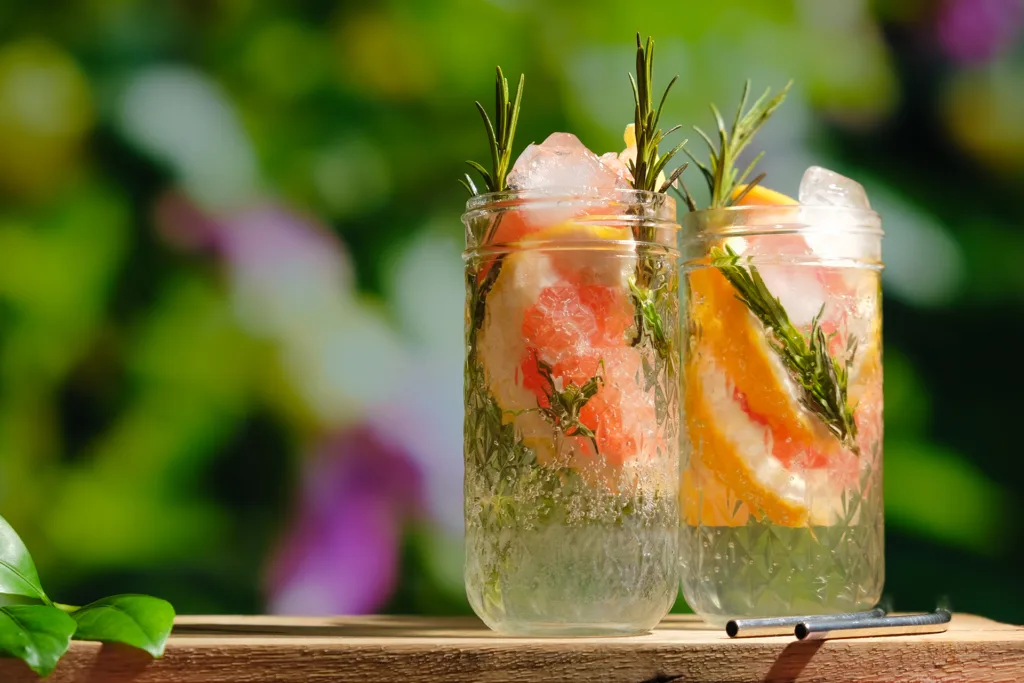 Deux verres d’eau aromatisée maison avec des tranches de pamplemousse, des branches de romarin et des glaçons sur fond estival