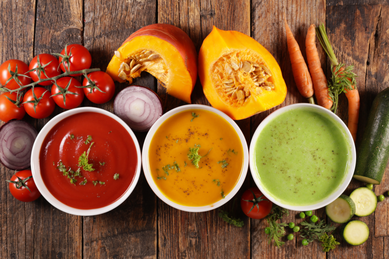 Trois bols de soupes maison aux légumes colorés : tomate, potimarron et courgettes, entourés d’ingrédients frais sur planche en bois rustique