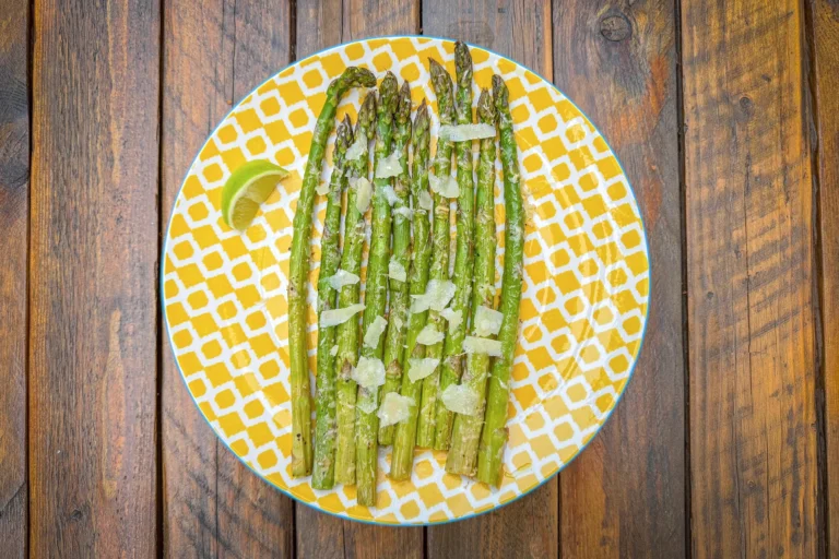 Assiette jaune de tiges d’asperges vertes rôties, copeaux de parmesan et quartier de citron vert surune table en bois