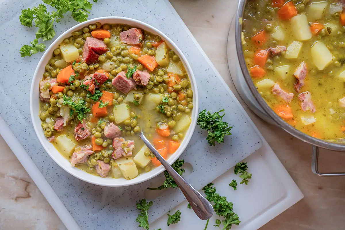 Bol de soupe paysanne aux petits pois, carottes, pommes de terre et morceaux de porc fumé sur planche blanche
