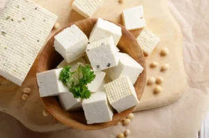 Cubes de tofu, un aliment plein de bienfaits, dans un bol en bois avec des graines de soja sur une planche.