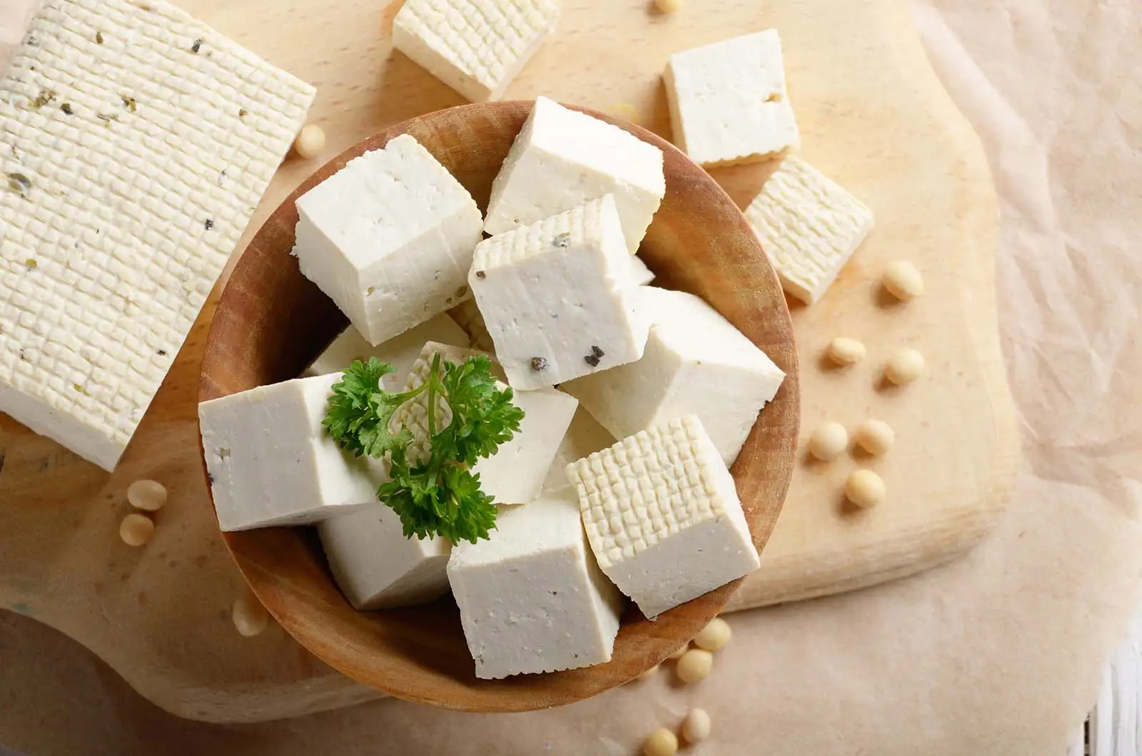 Cubes de tofu, un aliment plein de bienfaits, dans un bol en bois avec des graines de soja sur une planche.