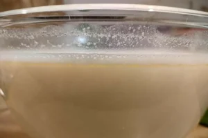 Pâte à cannelés bordelais dans un saladier en verre montrant une couche de mousse blanche d'air à la surface avant l'étape de l'écumage.