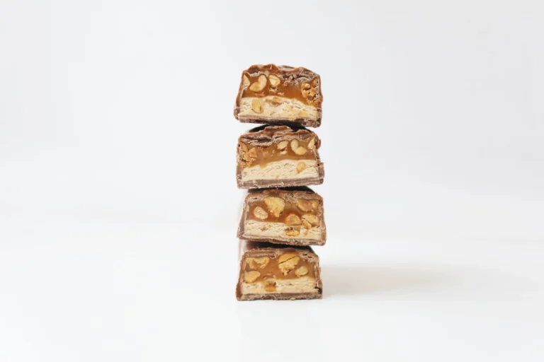Morceaux de barre chocolatée avec caramel, cacahuètes et nougat empilés sur fond blanc.