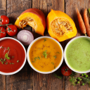 Trois bols de soupes maison aux légumes colorés : tomate, potimarron et courgettes, entourés d’ingrédients frais sur planche en bois rustique